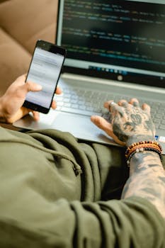 A focused individual coding on a laptop while checking a smartphone, embodying modern technology.