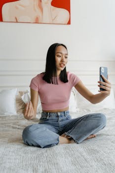 A young woman in casual attire takes a selfie while seated on a bed indoors.